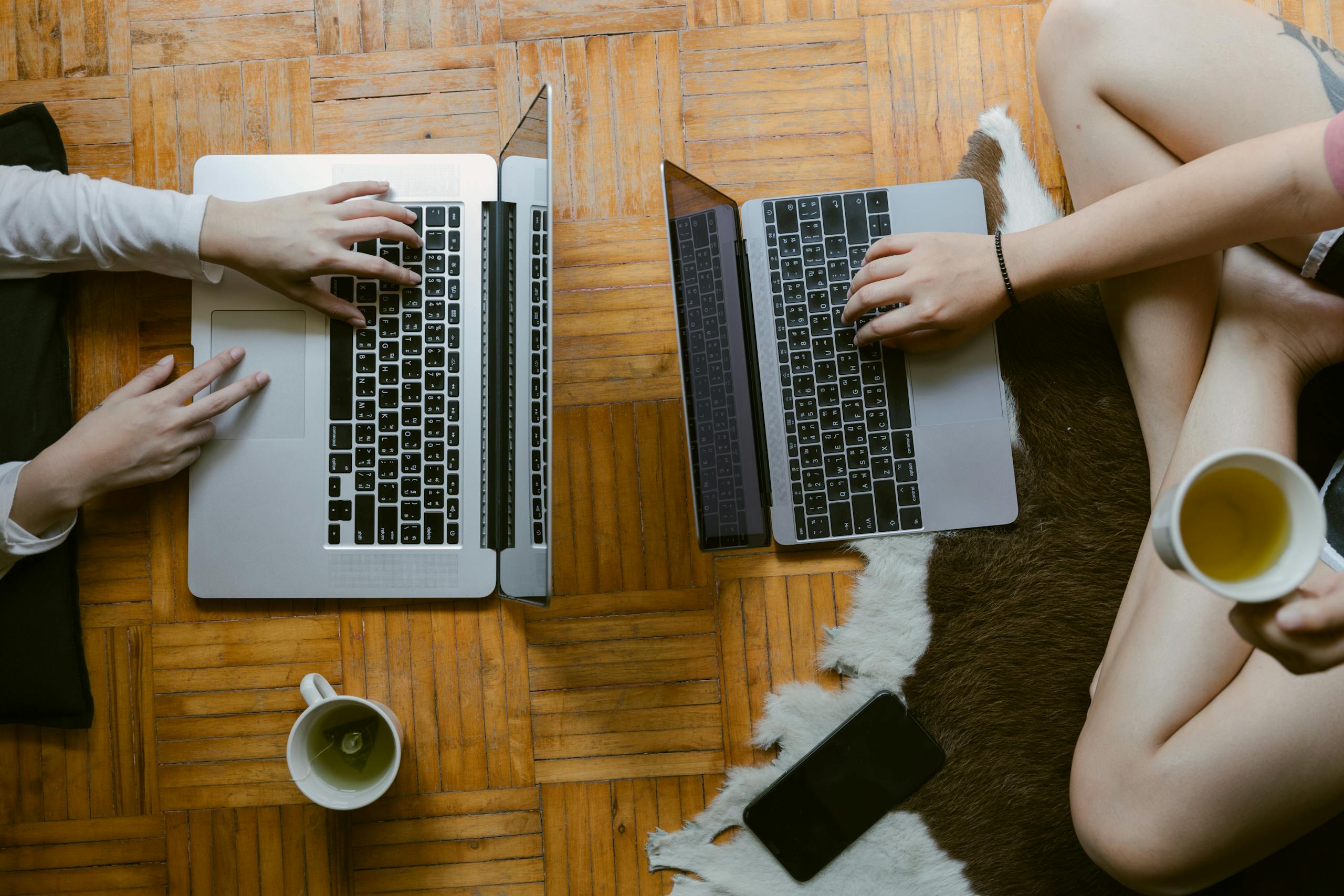 From above of crop unrecognisable female friends typing while studying on laptops remotely and enjoying tea on floor in cozy living room
