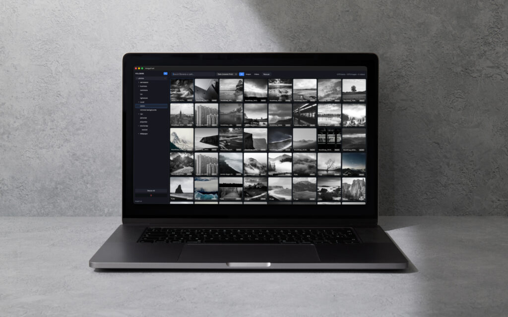 Laptop on a gray surface displaying a photo gallery grid of small grayscale thumbnails in a dark interface.