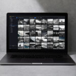 Laptop on a gray surface displaying a photo gallery grid of small grayscale thumbnails in a dark interface.