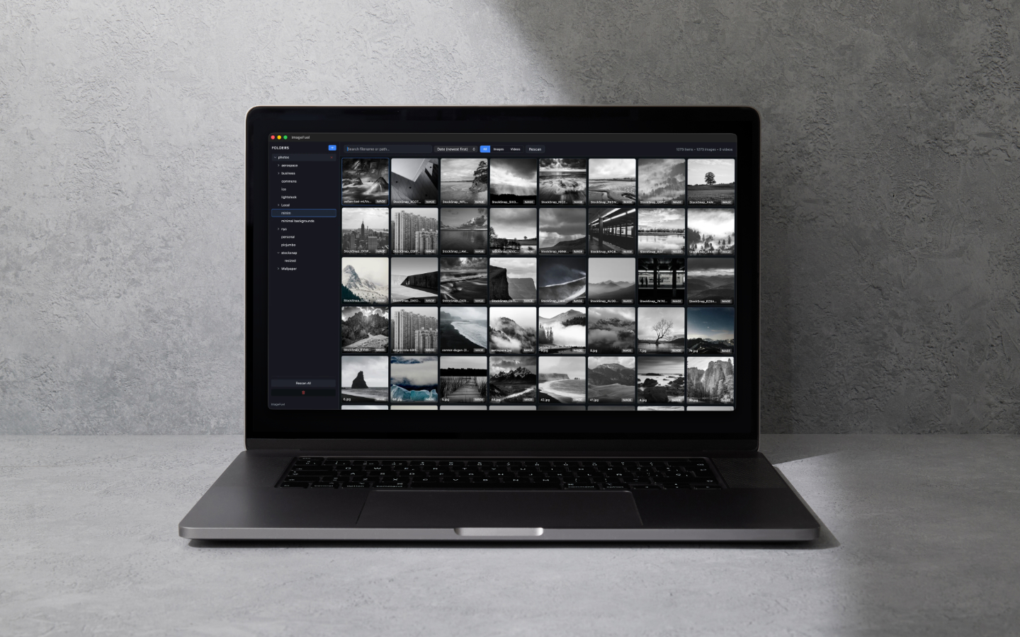 Laptop on a gray surface displaying a photo gallery grid of small grayscale thumbnails in a dark interface.